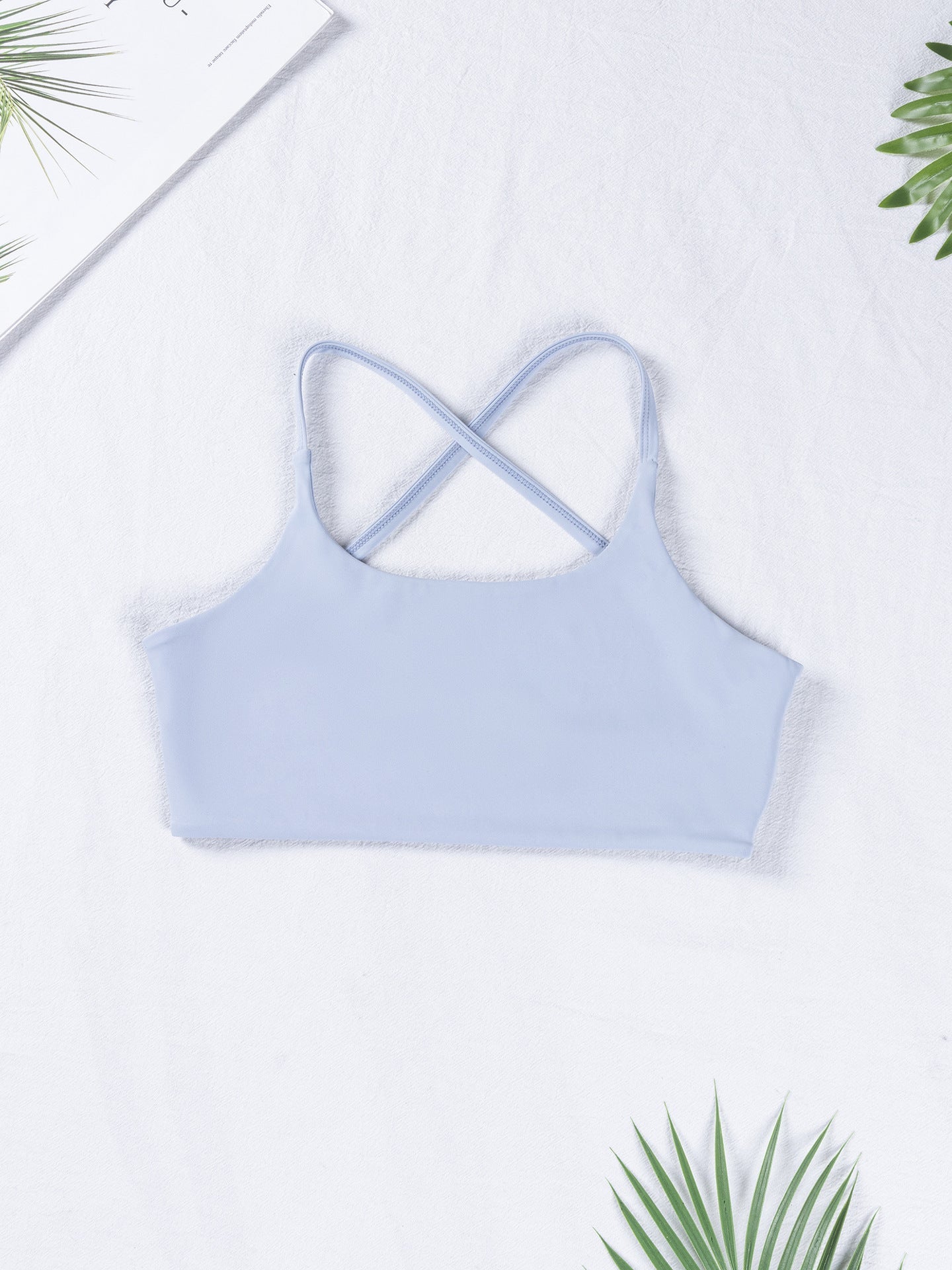 Light blue crop top with criss-cross straps on a white background with green palm leaves.