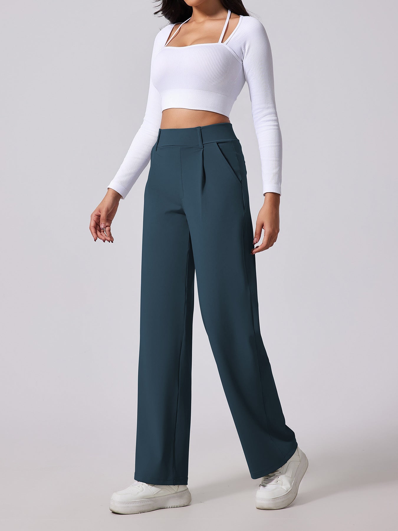 Person wearing a white long-sleeve top and teal high-waisted pants on a light gray background