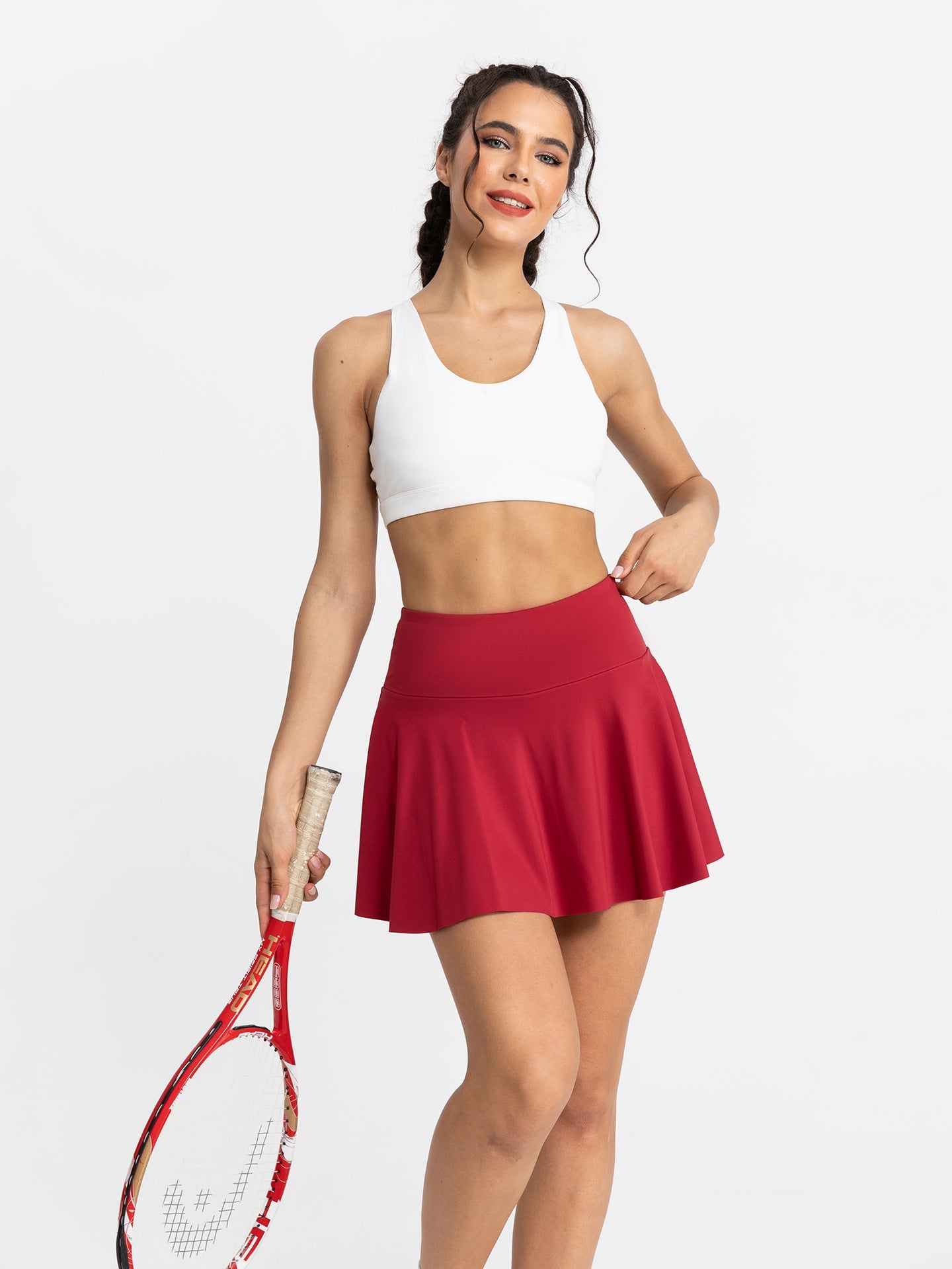 Woman in white sports bra and red tennis skirt holding a racket on a white background