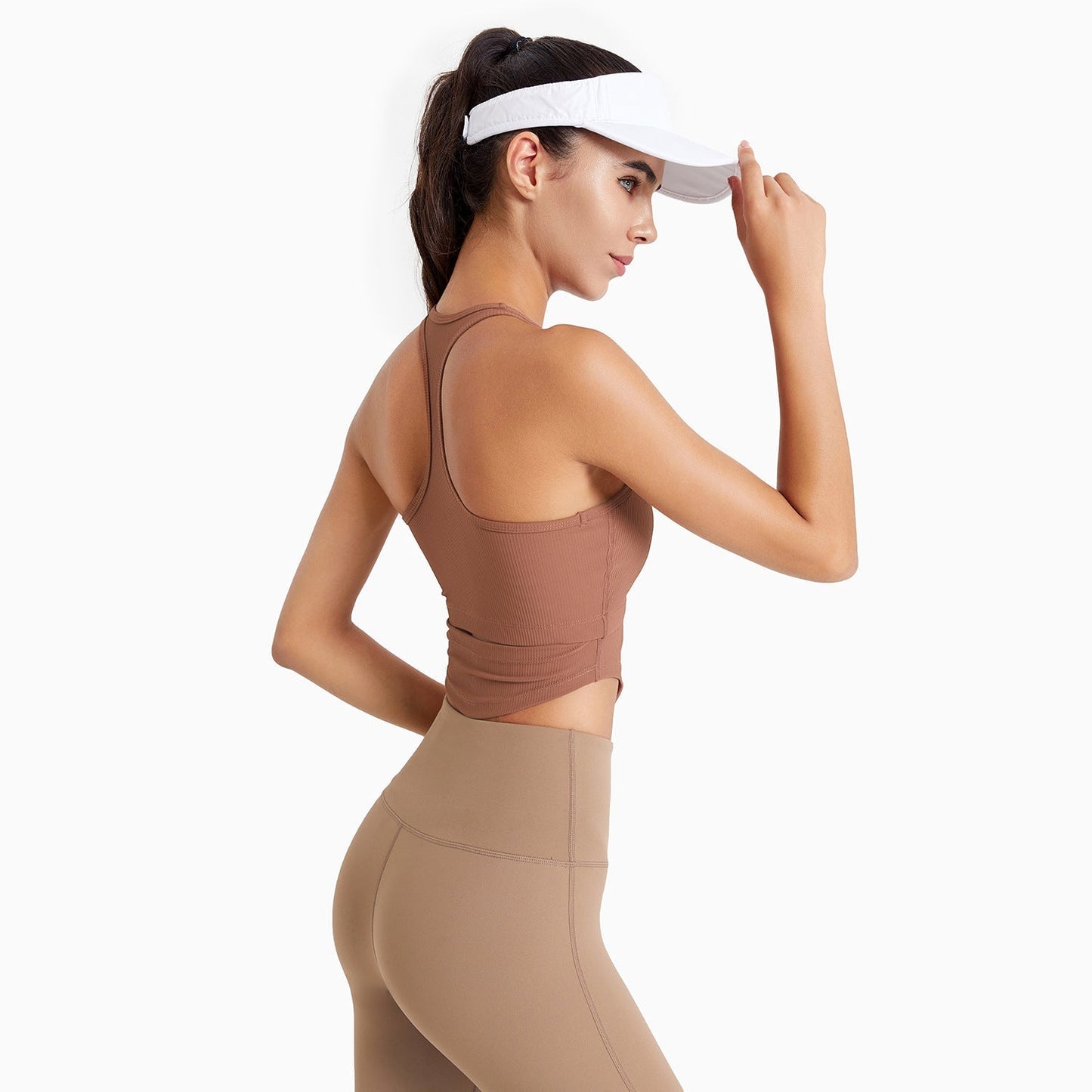 Woman wearing a brown sports bra and leggings with a white visor on a white background